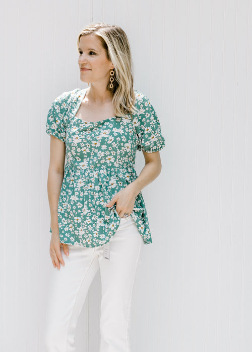 Model wearing white jeans with a green top with a white floral pattern and a square neckline. 