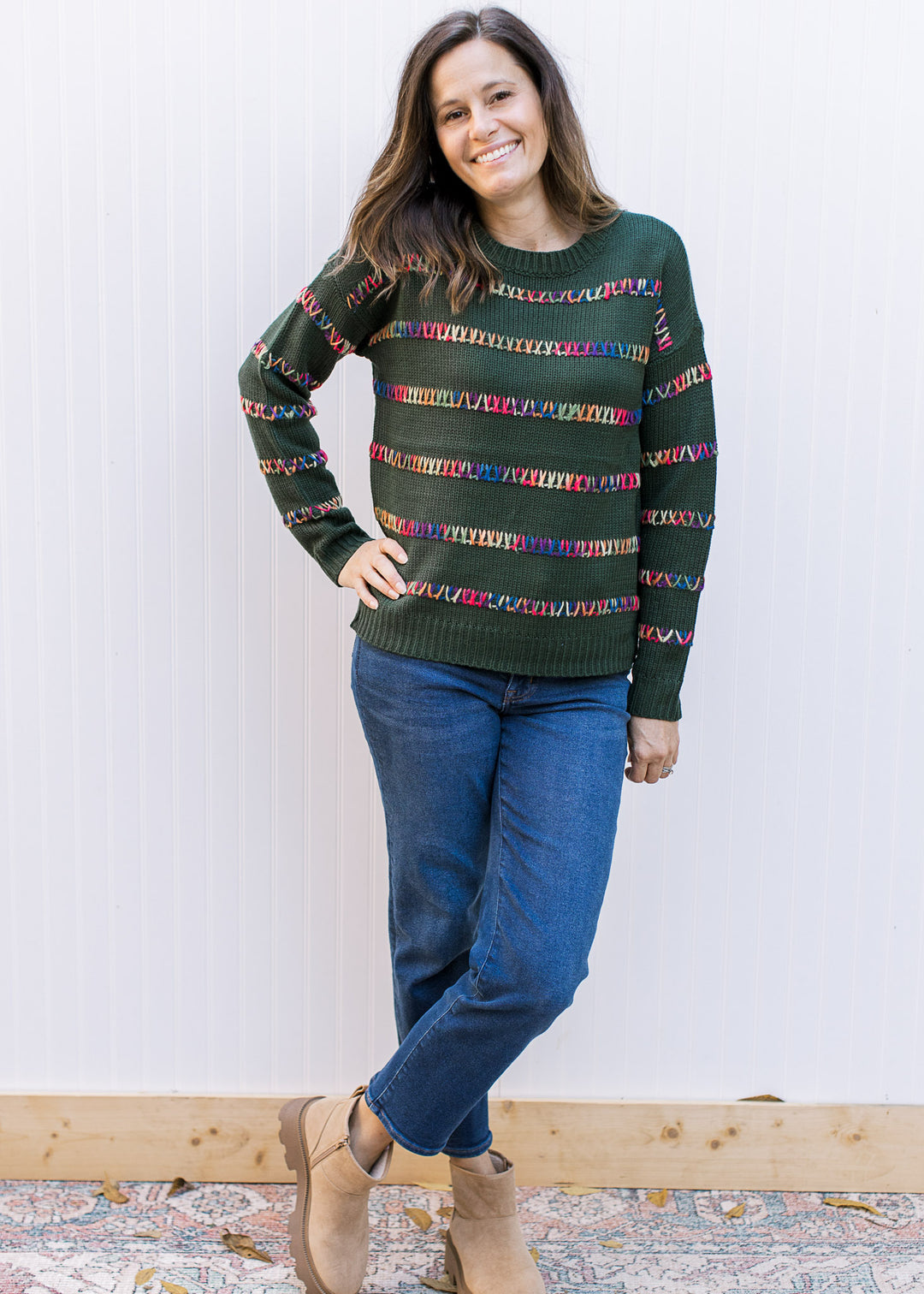 Model wearing jeans, booties and a hunter green sweater with colorfully stitched stripes. 