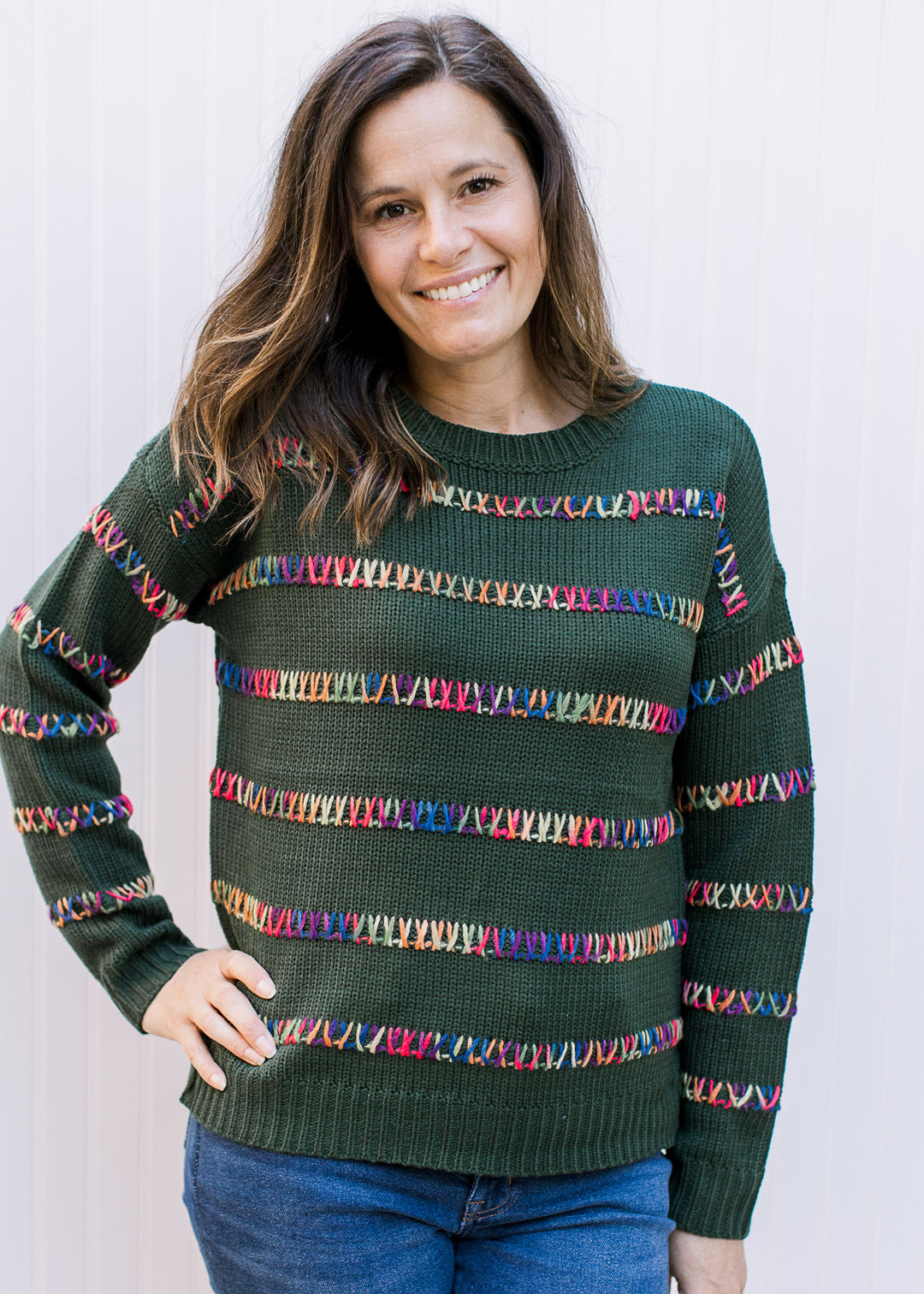 Model wearing a hunter green sweater with colorfully stitched stripes and long sleeves. 