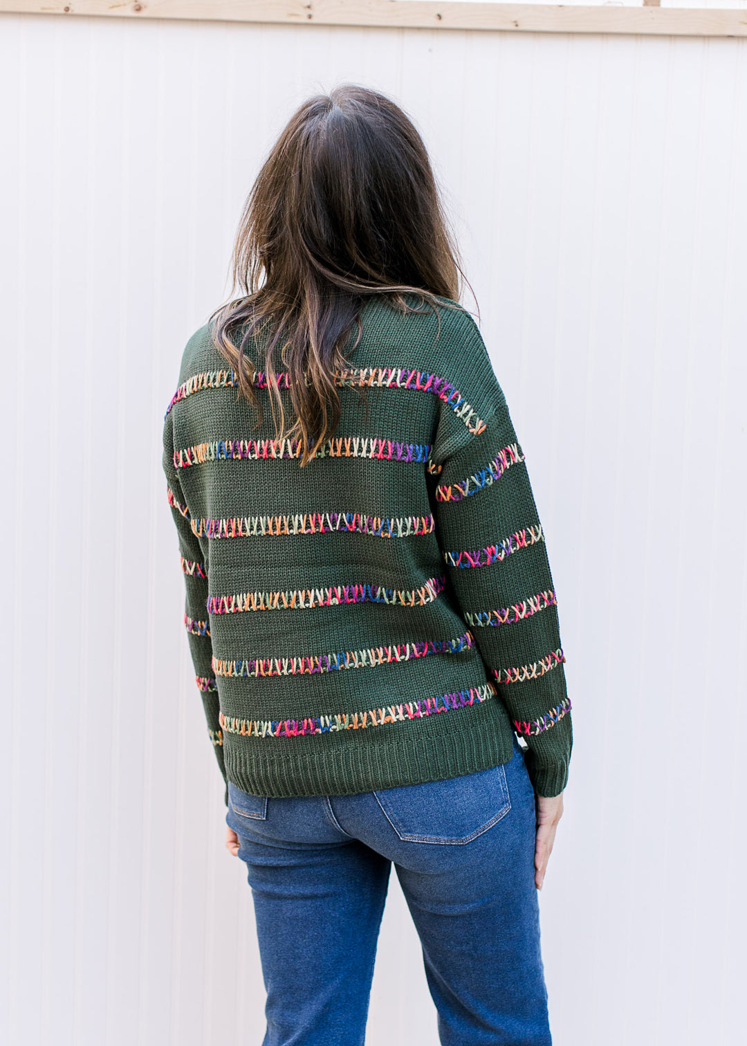 Back view of Model wearing a hunter green long sleeve sweater with colorfully stitched stripes. 