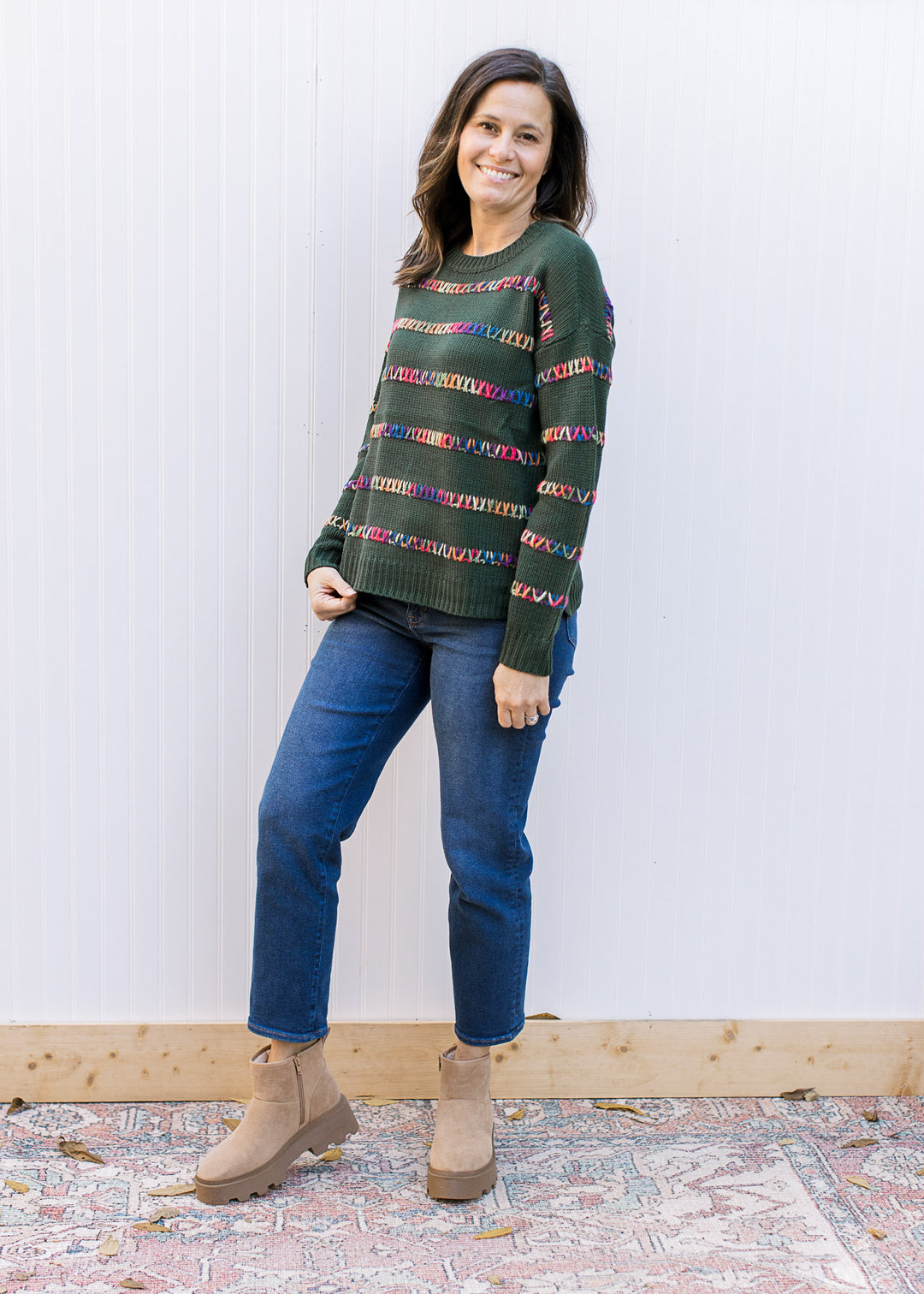 Model wearing jeans and a hunter green sweater with colorfully stitched stripes and long sleeves. 