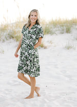 Model wearing an above the knee, button up black dress with a green and cream floral pattern. 