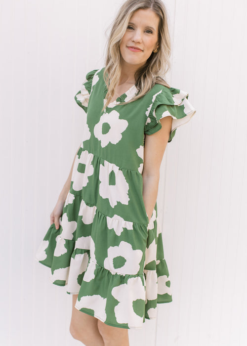 Model wearing a green dress with cream flowers, pockets and layered cap sleeves. 