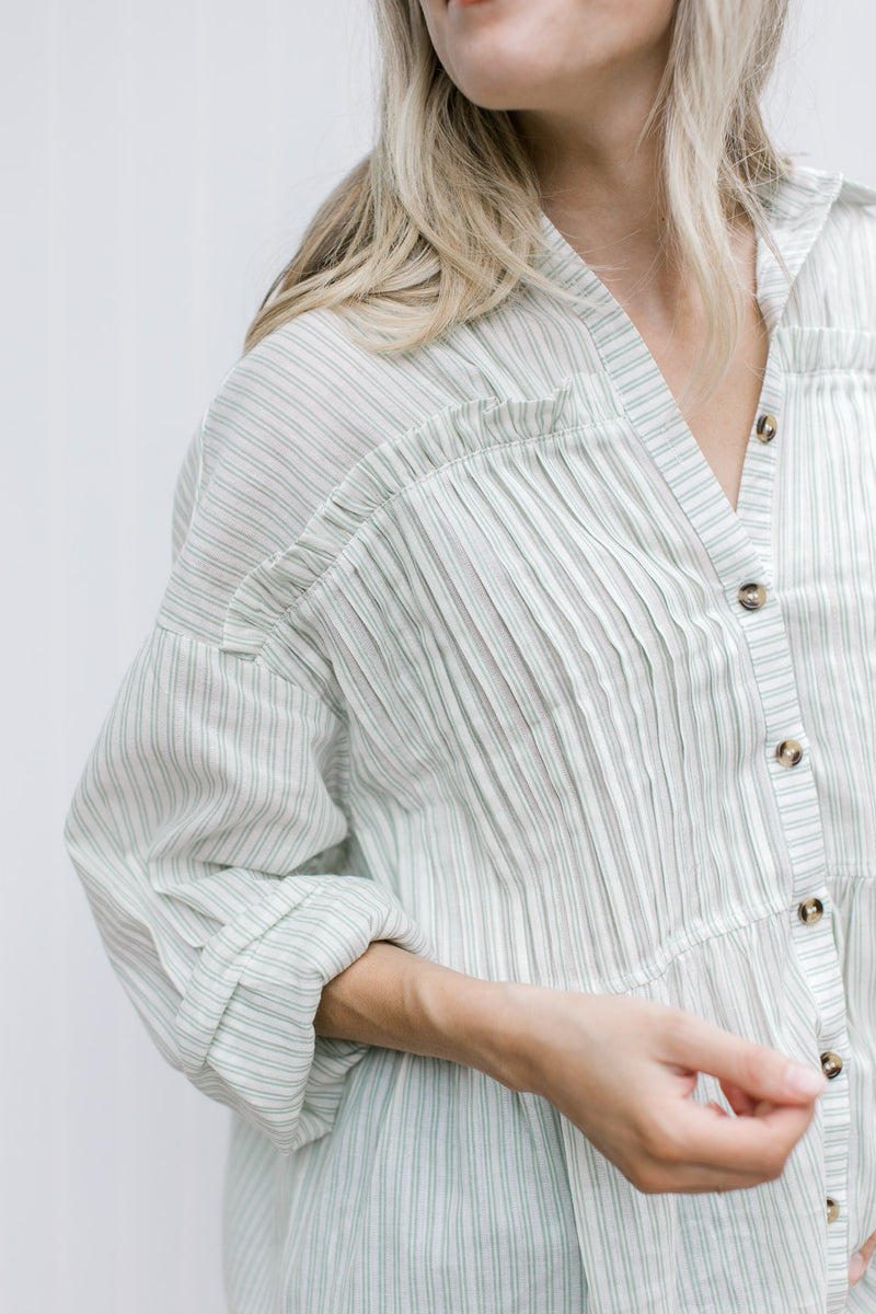 Close up of pleating on the front of a cream button up top with green pinstripes.