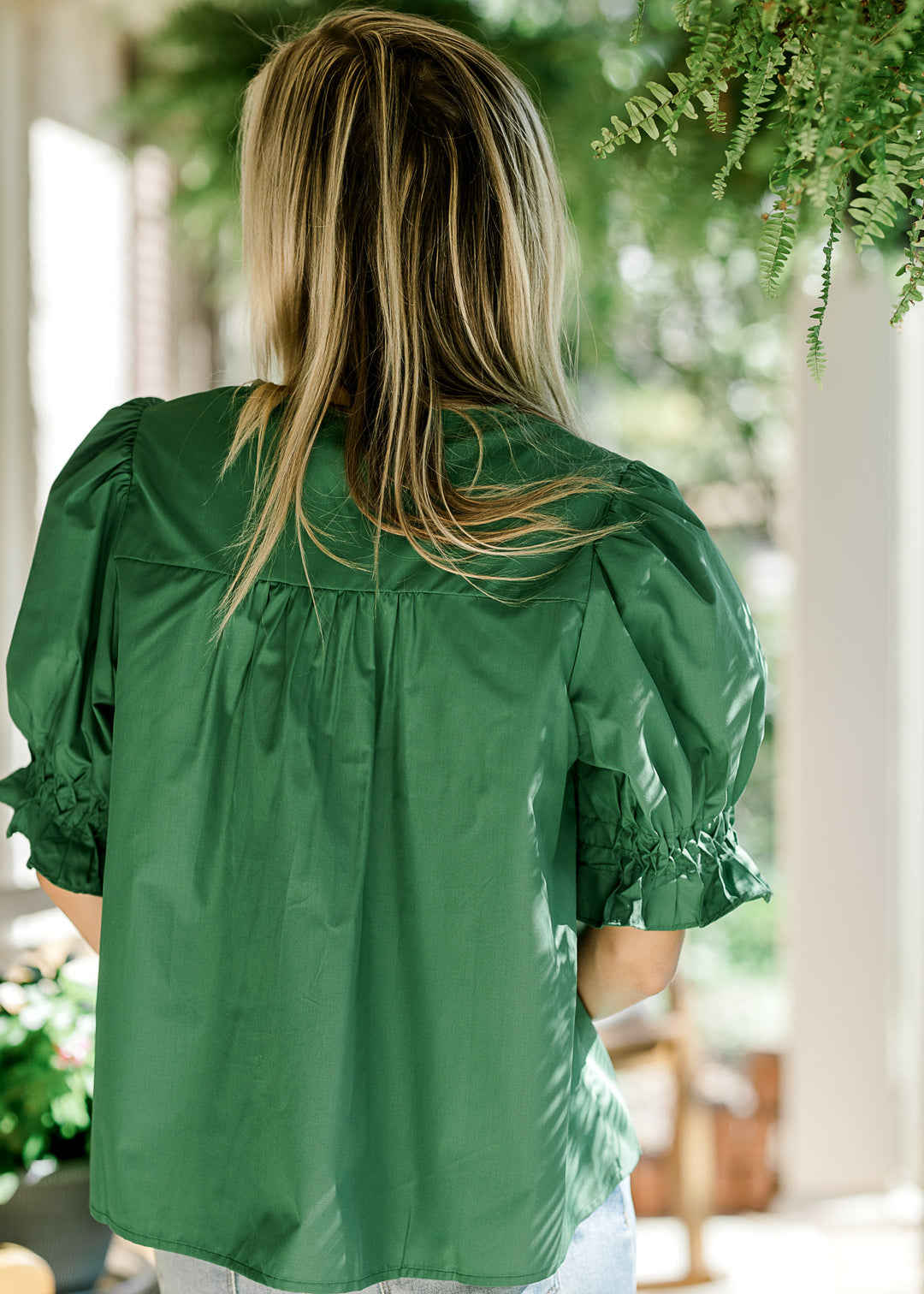 Back view of Model in a green blouse with a pleated front, bubble short sleeves and button front. 
