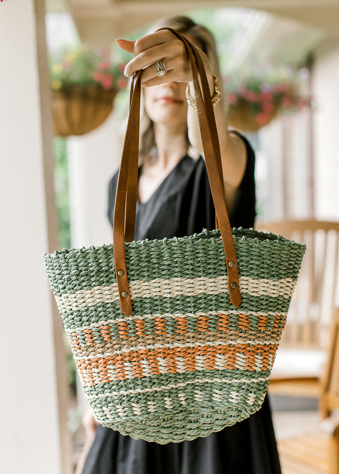 Model holding a woven straw purse with taupe, cream, green, and rust pattern a faux leather straps.