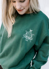 Model wearing a deep green sweatshirt with a round neck and an embroidered white golf club logo. 