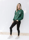 Model wearing black leggings and a deep green sweatshirt with a round neck and an embroidered logo. 