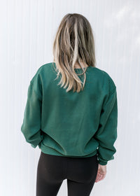 Back view of Model wearing a deep green sweatshirt with a round neck and an embroidered lego.