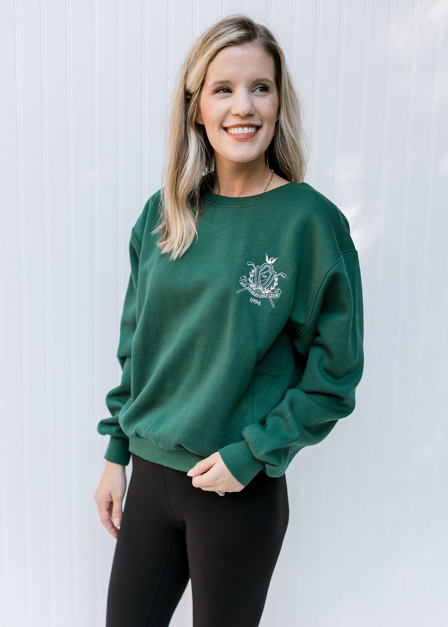 Model wearing a deep green sweatshirt with a round neck and an embroidered white logo. 