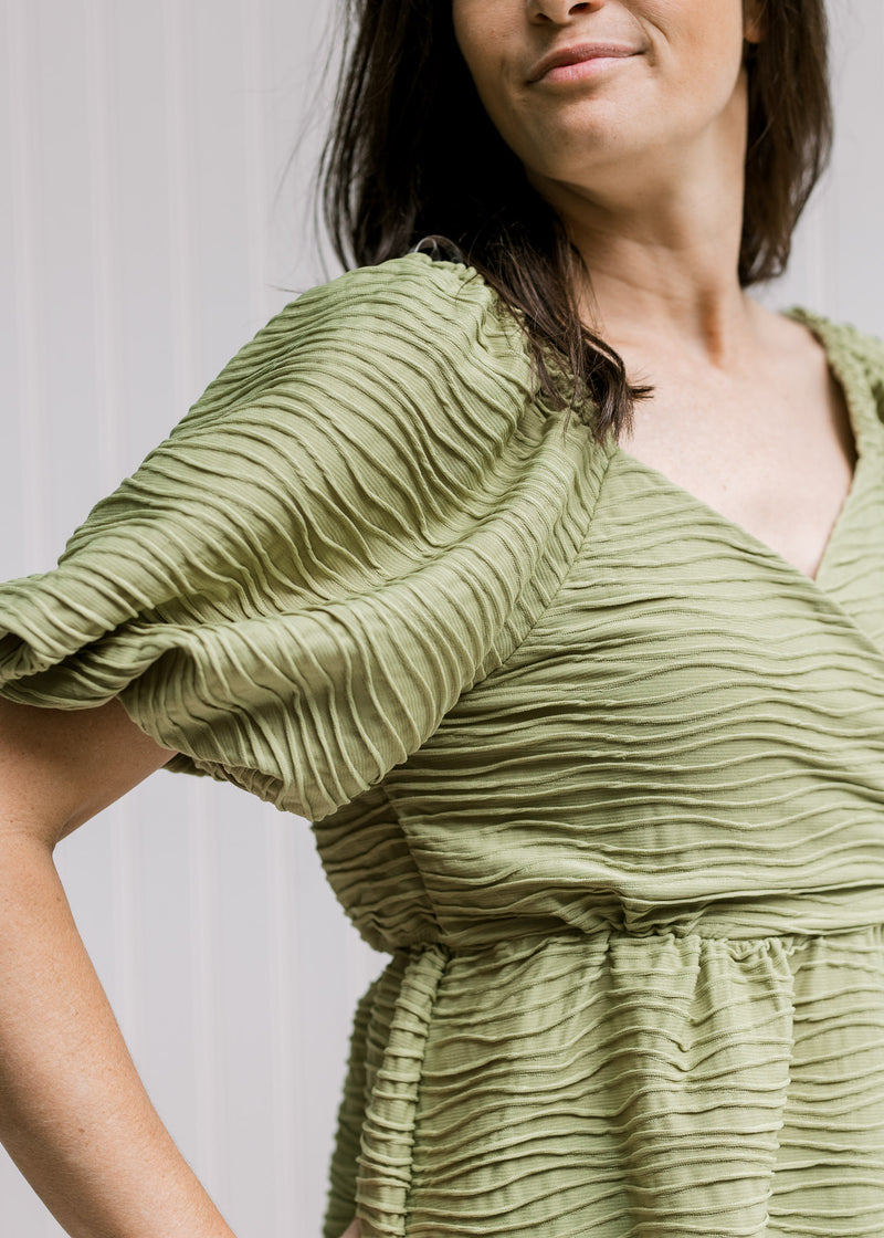 Close up of v-neck with snap, elastic waist and bubble short sleeves on an avocado colored top. 