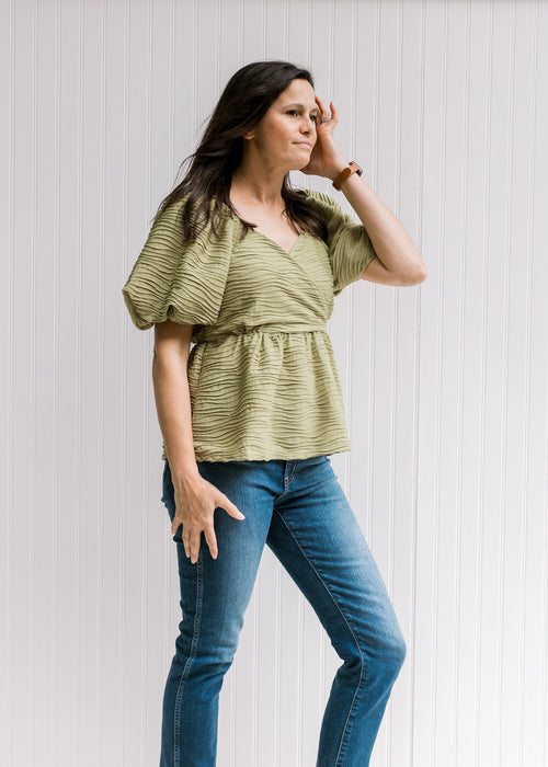 Model wearing a avocado green v-neck top with a pleated wave texture and short bubble sleeves. 
