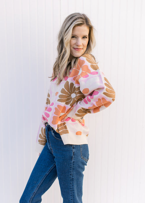 Model wearing a cream sweater with oversized orange, pink, rust and taupe flowers and long sleeves.