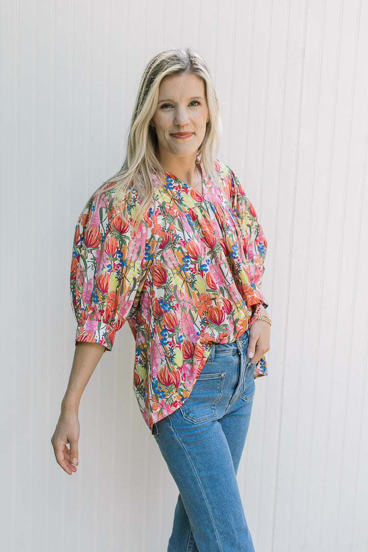 Model wearing jeans with a white top with bright floral patter, 3/4 sleeve with cuff and v-neck.