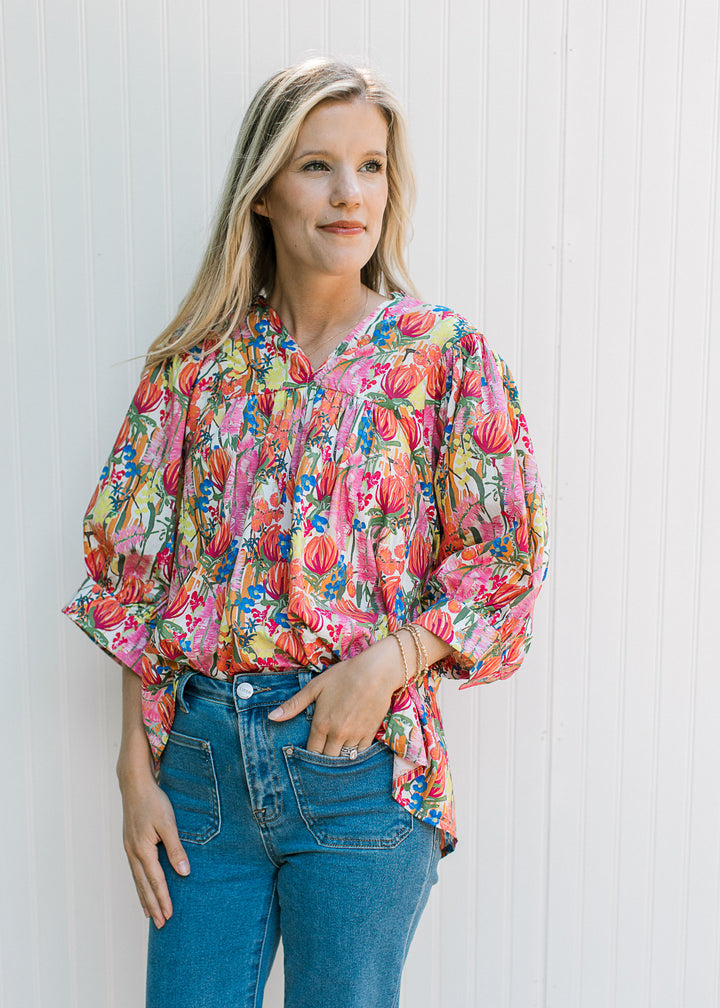 Model wearing a white top with bright floral patter, 3/4 sleeve with cuff and ruffle at neck.