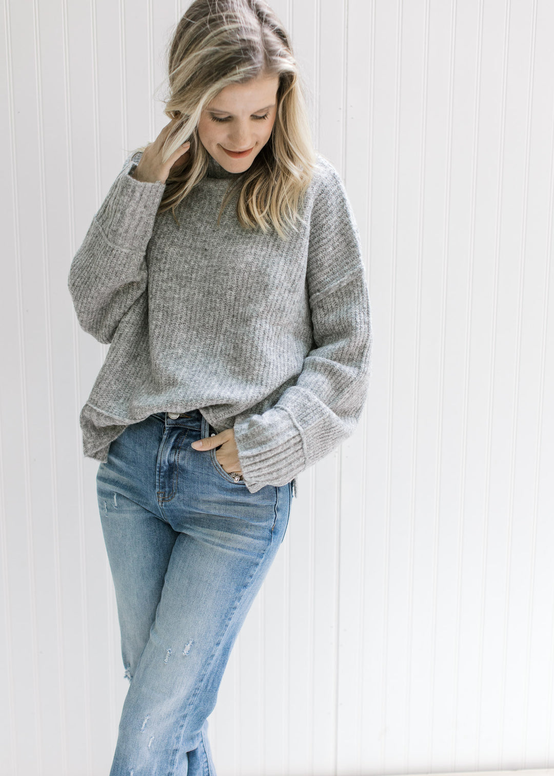 Model wearing a gray ribbed sweater with exposed hem, long sleeves and a mock neckline. 