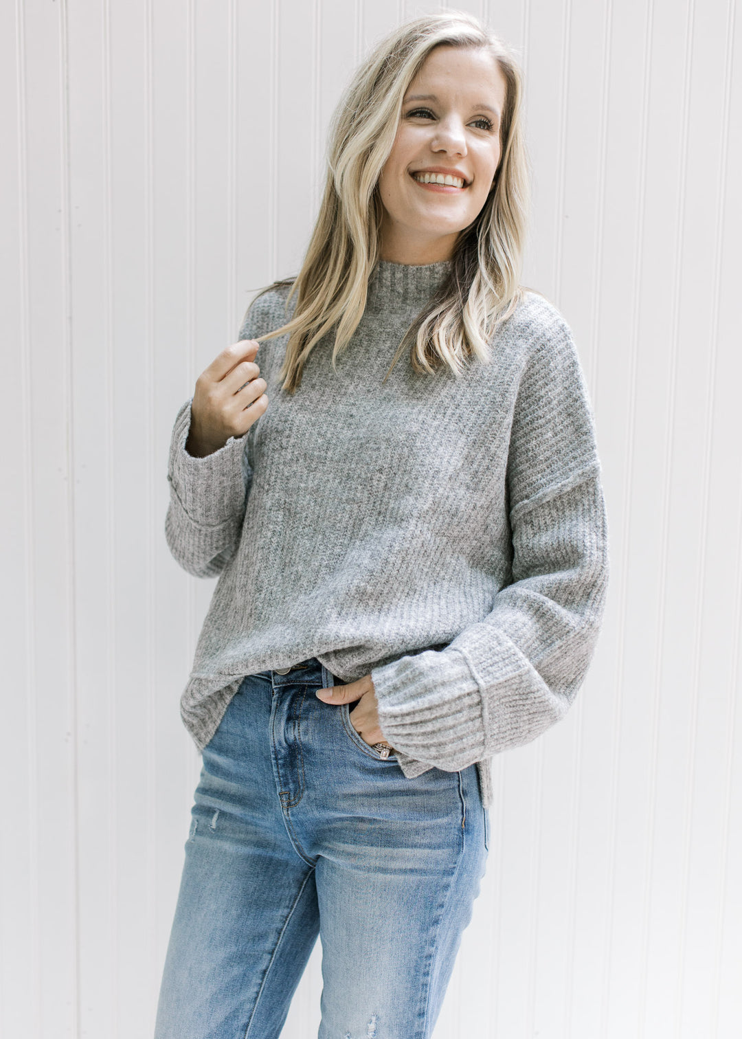 Model wearing a gray ribbed sweater with exposed hem, long sleeves and a mocked neck. 