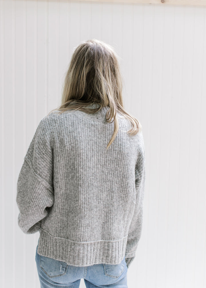 Back view of Model wearing a gray ribbed sweater with exposed hem, long sleeves and a mock neck. 