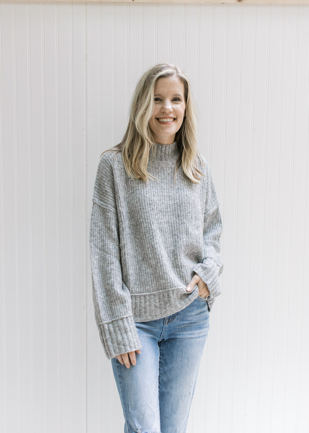 Model wearing jeans with a gray ribbed sweater with exposed hem, long sleeves and a mock neckline. 