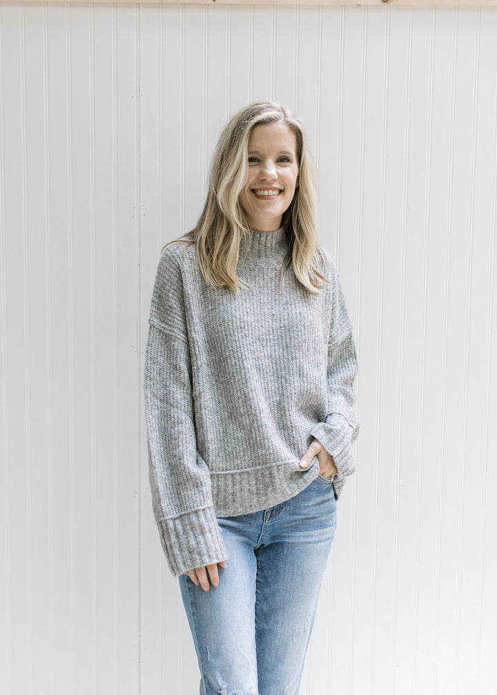 Model wearing jeans with a gray ribbed sweater with exposed hem, long sleeves and a mock neckline. 