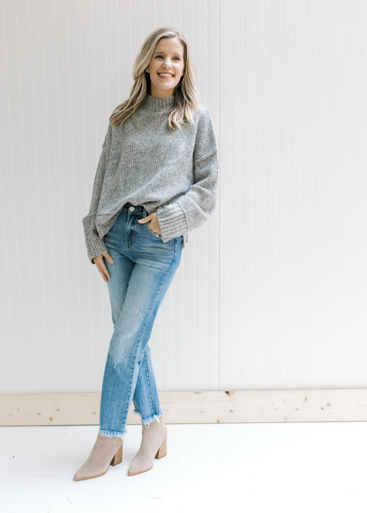 Model wearing jeans, booties and a gray sweater with exposed hem, long sleeves and a mock neck. 