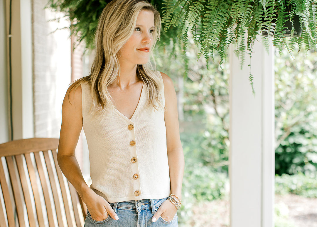 Model wearing a sleeveless, slightly cropped cream knit sweater with a v-neck and button front. 