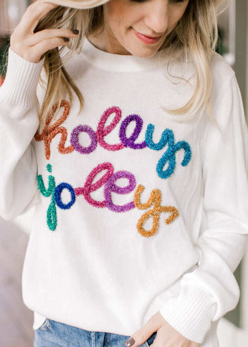 Model wearing a white sweater with the words holly jolly across the front in multicolored tinsel. 