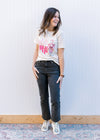 Model wearing jeans, sneakers and a cream short sleeve tee with holly jolly in pink text.