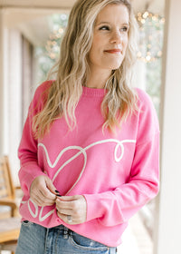 Model wearing a hot pink sweater with a cream heart design along the front and long sleeves. 