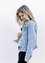 Side view of Model wearing a light denim jacket with a button closure, raw hem and long sleeves. 