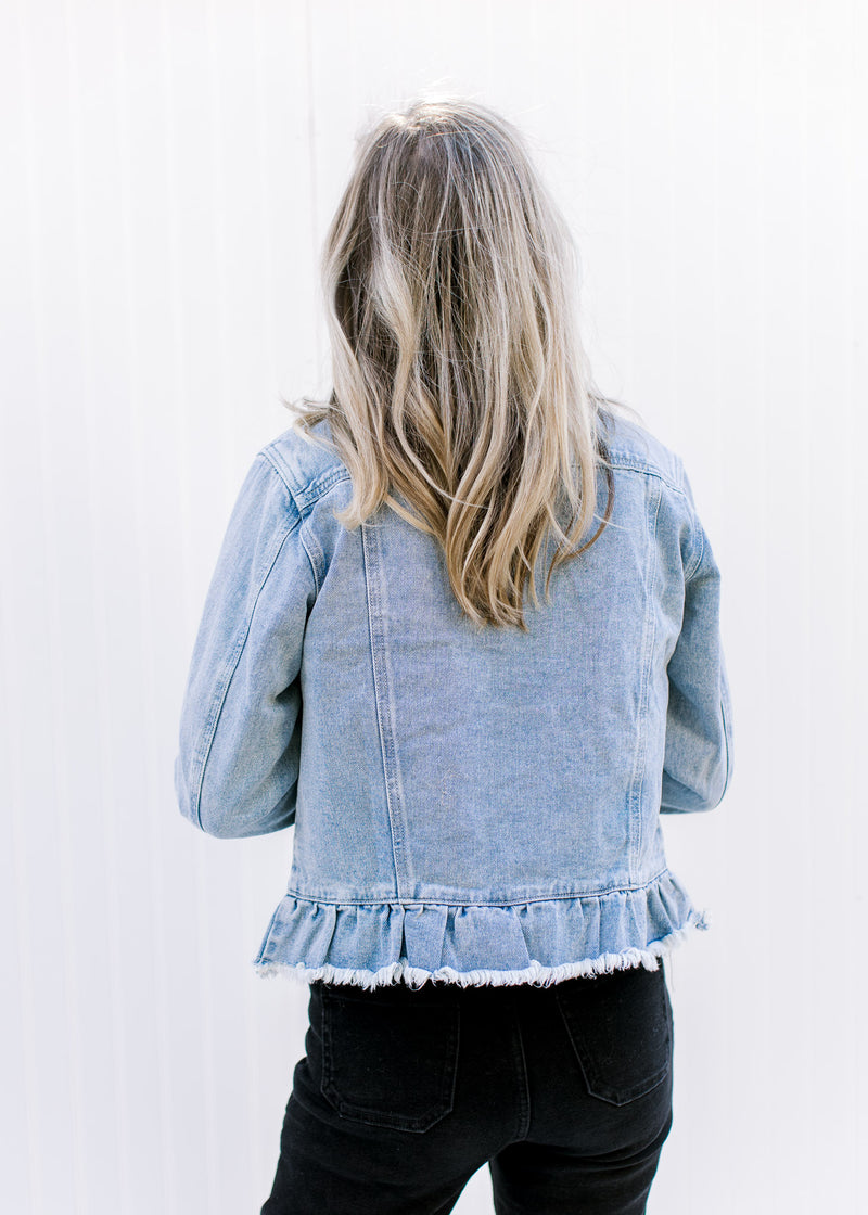 Back view of Model wearing a light denim jacket with a button closure, raw hem and long sleeves. 