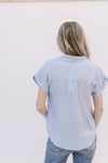 Back view of Model wearing a pale blue top with patch pockets, collar and rolled short sleeves.