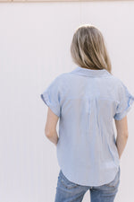 Back view of Model wearing a pale blue top with patch pockets, collar and rolled short sleeves.