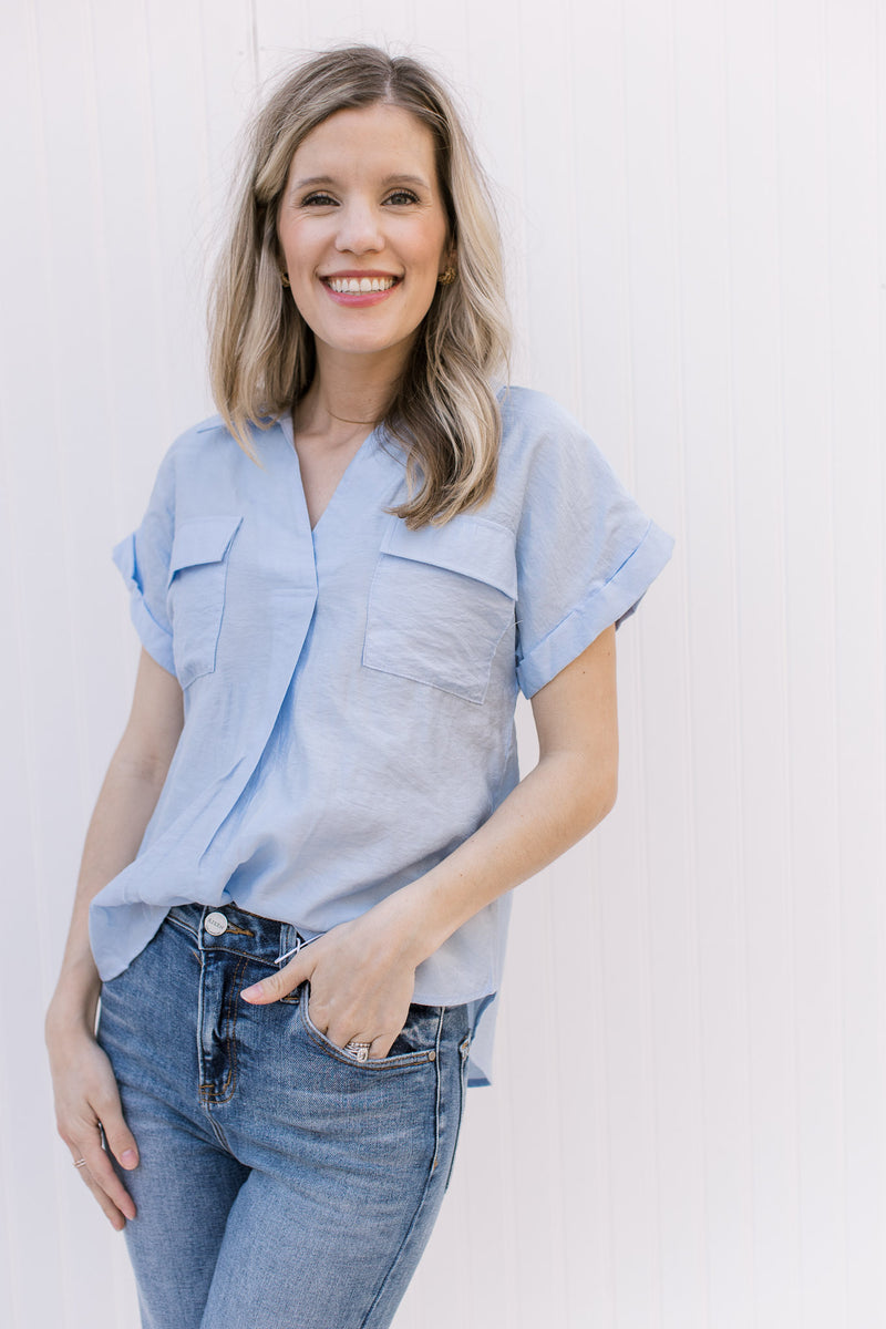 Model wearing jeans and a pale blue top with patch pockets, v-neck, collar and rolled short sleeves.