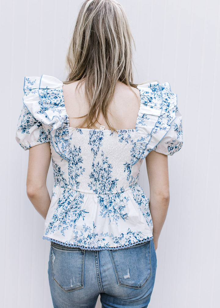 Back view of Model wearing a white short sleeve top with blue floral, square neck and smocked back.