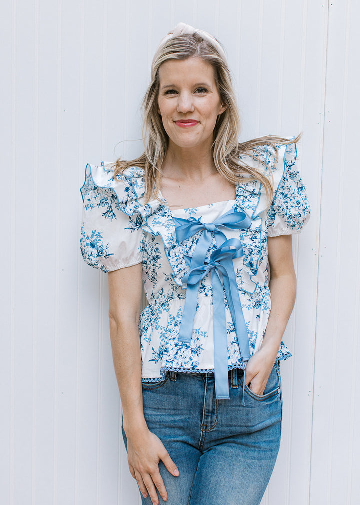 Model wearing a white short sleeve top with blue floral, ruffle at shoulders and double bow ties.