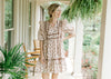 Model wearing a cream dress with taupe and brown flowers, ruffle at shoulder and short puff sleeves.