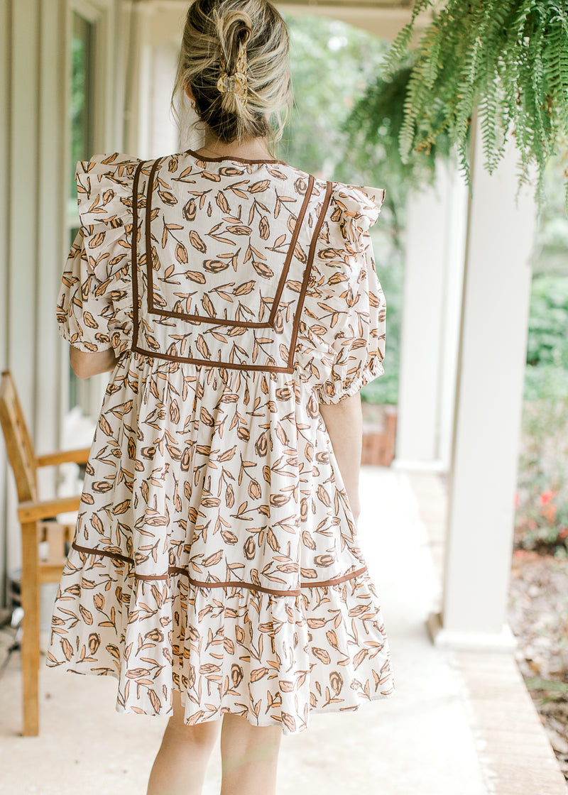 Back view of Model wearing a cream dress with taupe and brown flowers and short puff sleeves.