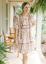 Model wearing a cream dress with taupe and brown flowers, v-neck with tie and short sleeves.