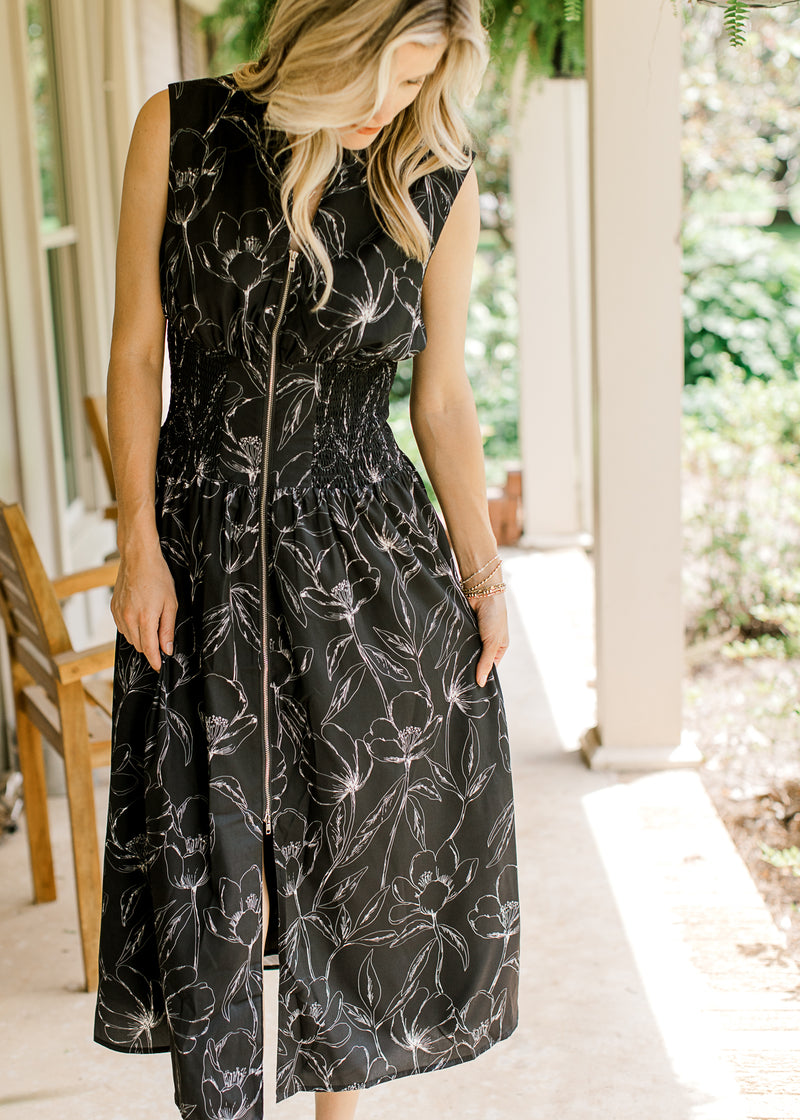 Model wearing a black sleeveless midi with white floral, smocked waist and full zip. 