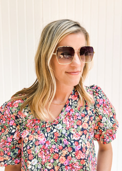 Model wearing sunglasses with a gold wire frame and gray ombré lenses with a floral top. 