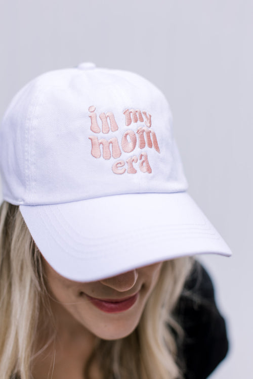 Model wearing an white adjustable size hat with pink embroidered script “in my mom era”. 