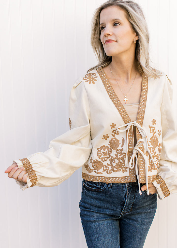 Model wearing a slightly cropped khaki jacket with tan floral, lace detailing and a two tie closure.