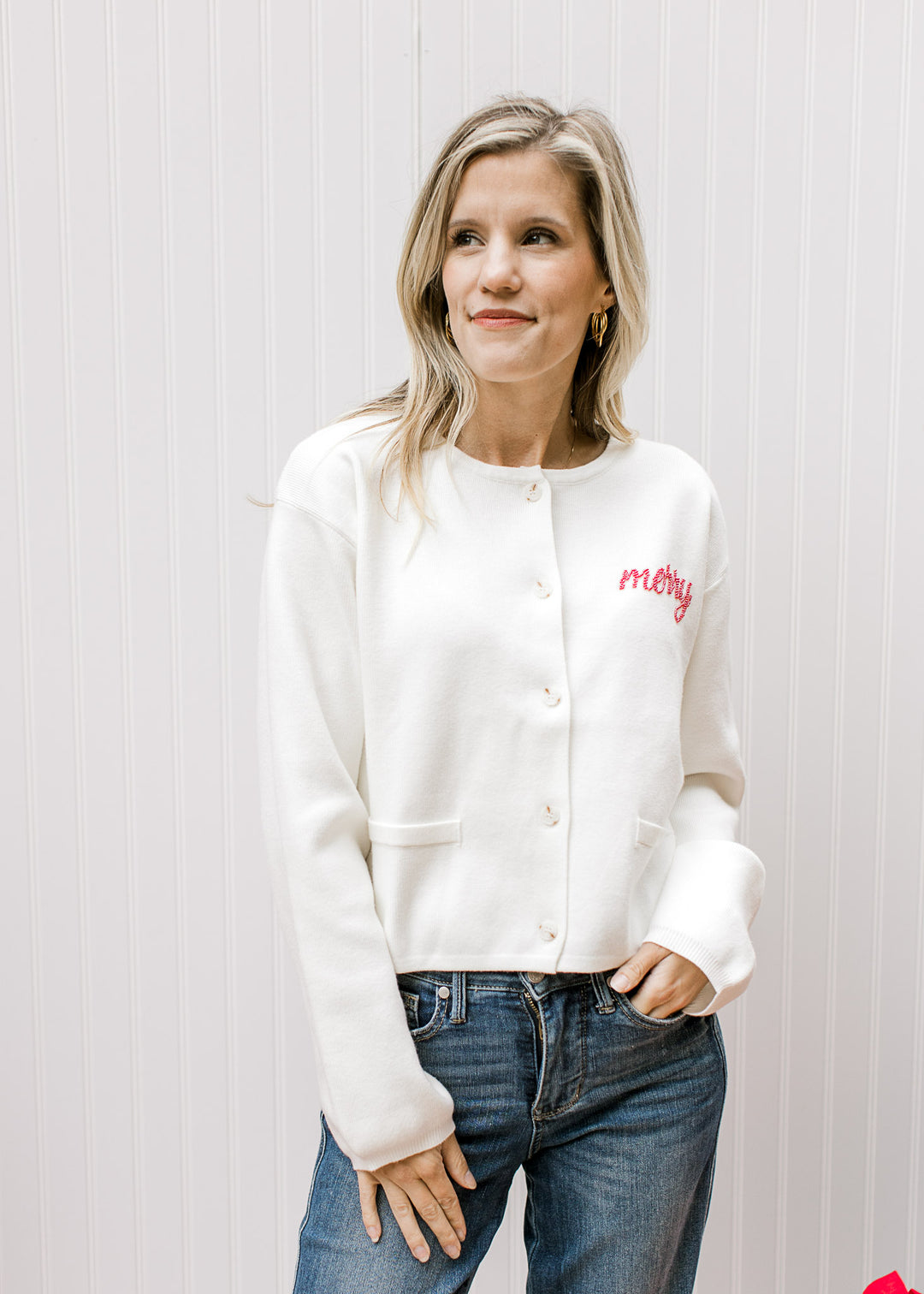 Model wearing a slightly cropped ivory cardigan with “Merry” written in red beaded script. 
