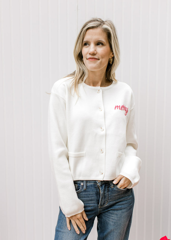 Model wearing a slightly cropped ivory cardigan with “Merry” written in red beaded script. 
