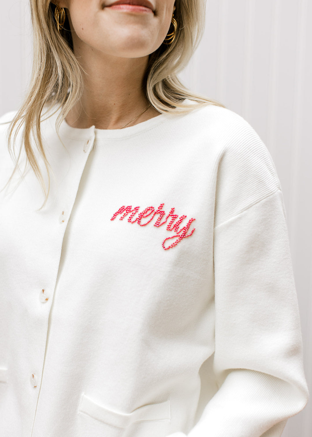 Model wearing a long sleeve ivory cardigan with “Merry” written in red beaded script at left chest. 