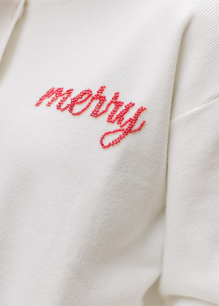 Close up of scripted “merry” written in red beads on a cream ribbed long sleeve cardigan. 