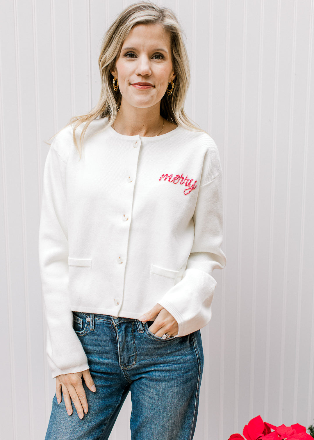 Model wearing an ivory cardigan with “Merry” written in red beaded script and front slit pockets. 