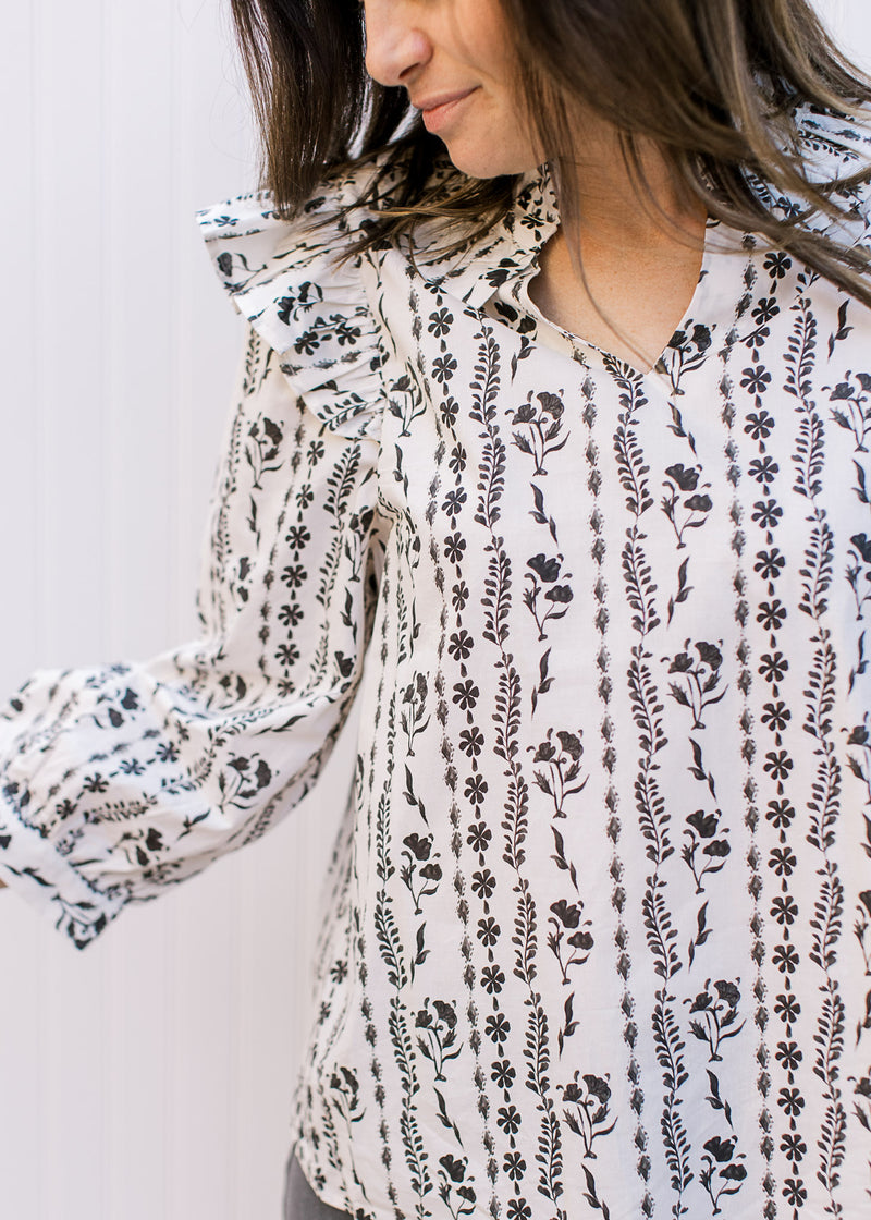 Model wearing a white top with black floral stripes with ruffle at shoulder and a v-neck. 