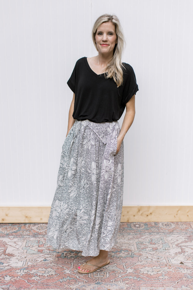 Model wearing a black top, sandals and an ivory skirt with black flowers and pockets. 